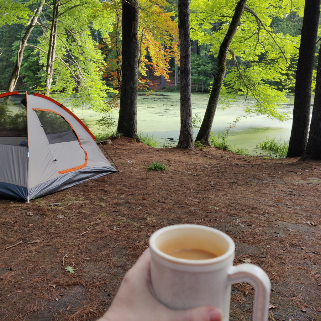 It's the little things that keep us going when everything feels too big. An image depicting a hand holding a full mug of coffee. In the background is an orange and light grey 2-person tent, several trees beginning to take on fall colors, and a pond covered with duckweed.